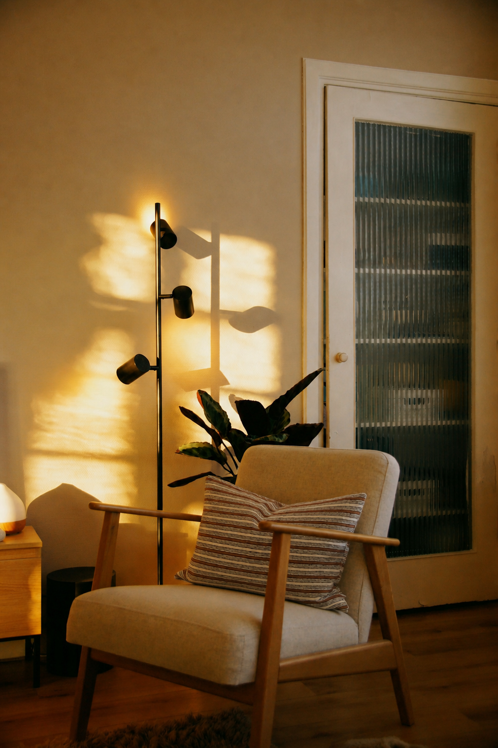 Poltrona in tessuto beige con cuscino Dino a righe nei toni naturali, illuminata da luce calda, casa con atmosfera rilassante, con lampada da terra, pianta verde e atmosfera intima e accogliente.