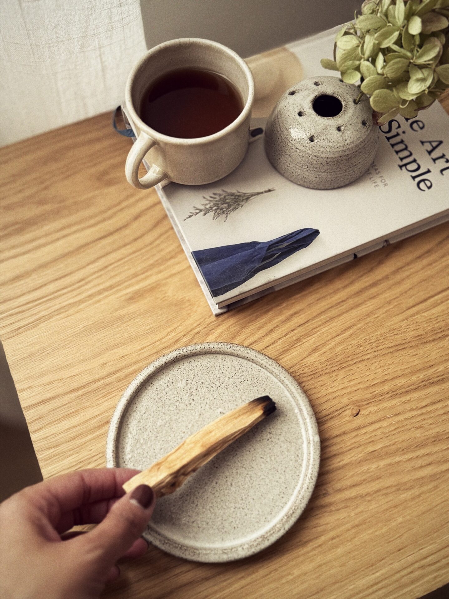 Una mano appoggia il palo santo sul piattino in gres, accanto a una tazza e a un vaso portafiori.