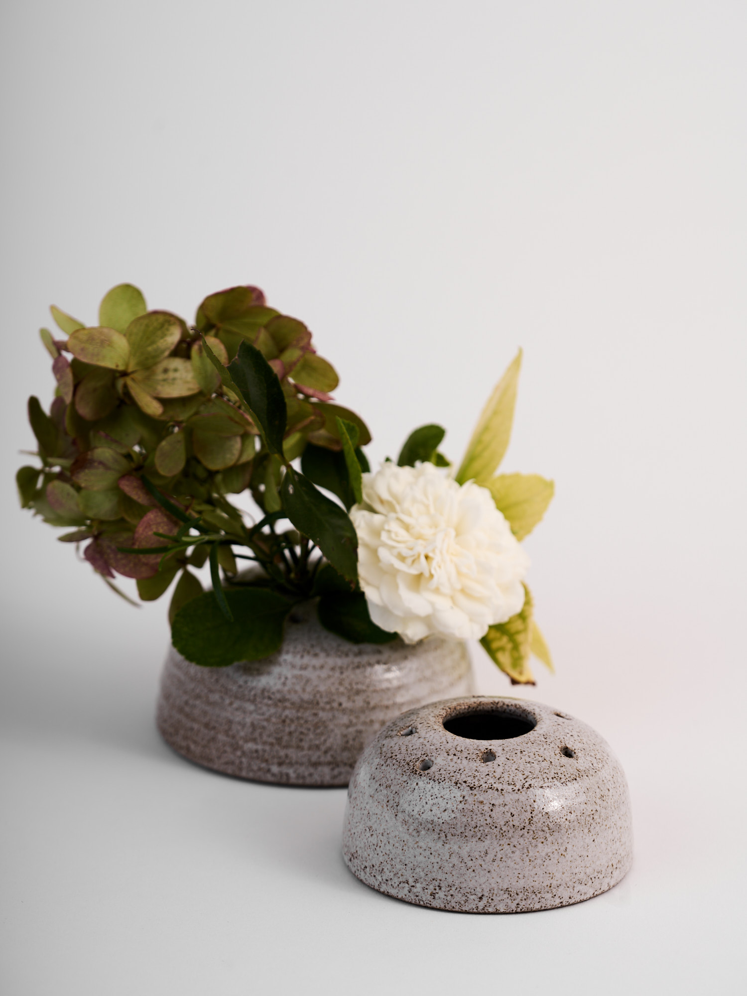 Due vasi portafiori in gres puntinato, uno vuoto e uno con fiori bianchi e verdi.